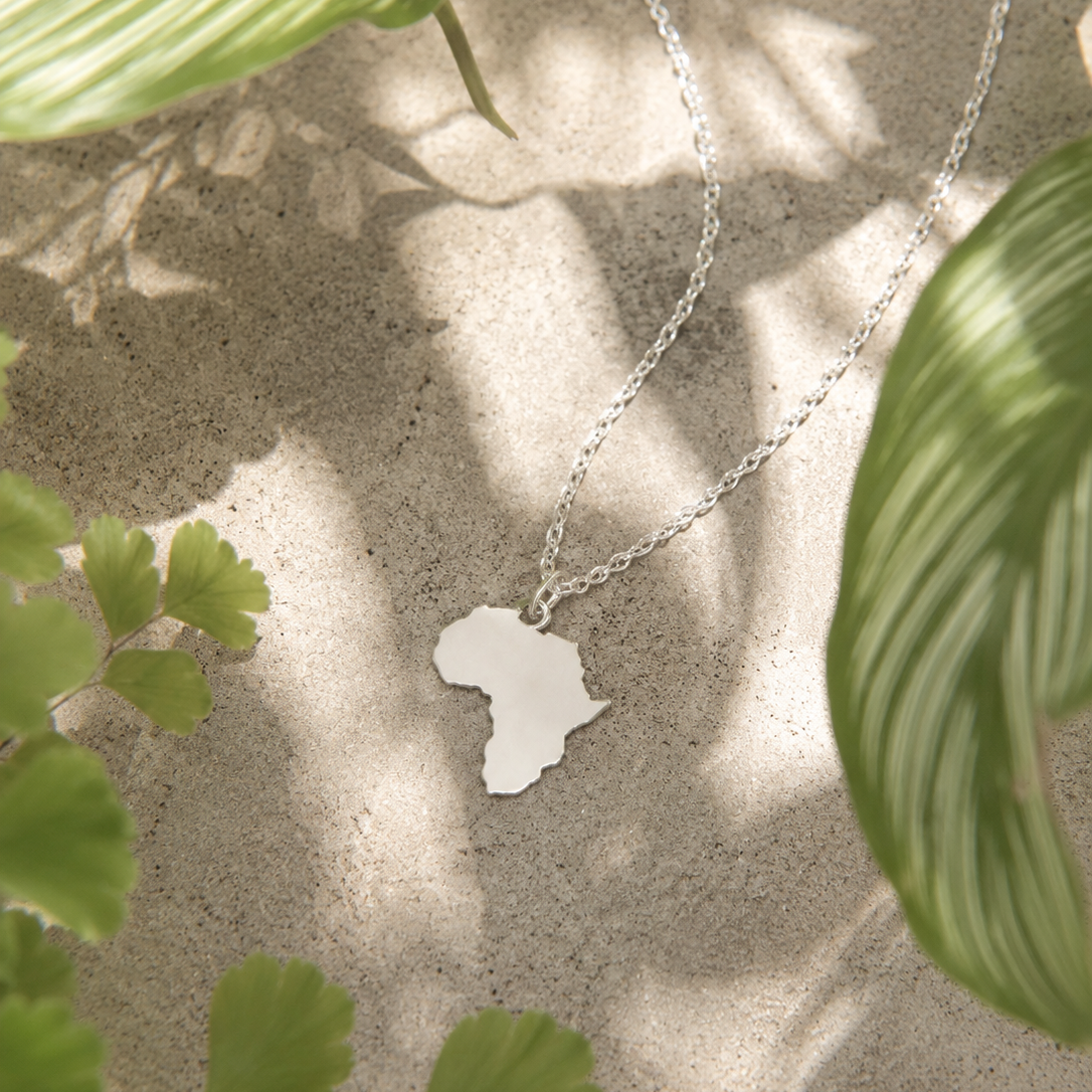 Silver necklace with a pendant shaped like Africa, surrounded by green leaves on a textured surface.