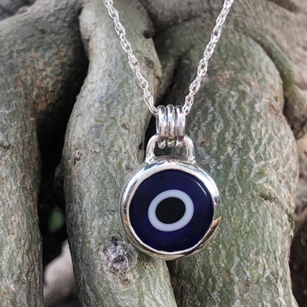 Handcrafted sterling silver evil eye necklace with a blue lampwork bead displayed on a tree trunk in natural outdoor light.