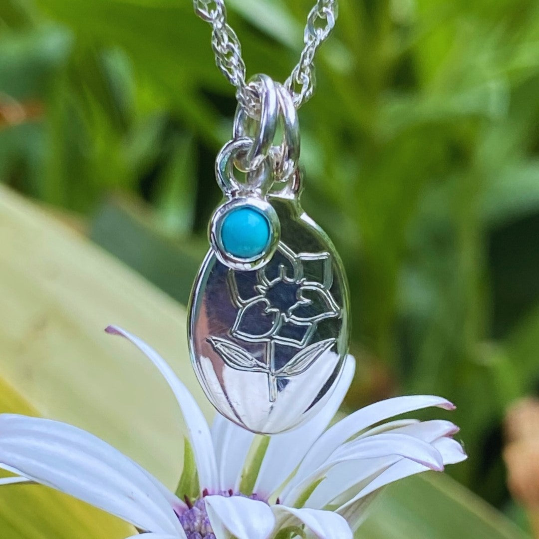 Handcrafted sterling silver birth flower necklace with turquoise gemstone displayed on a white flower.