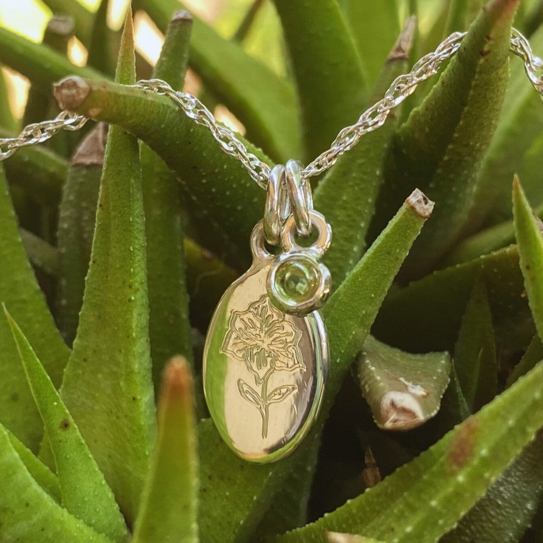 Handcrafted sterling silver birth flower necklace with gemstone displayed among green foliage.